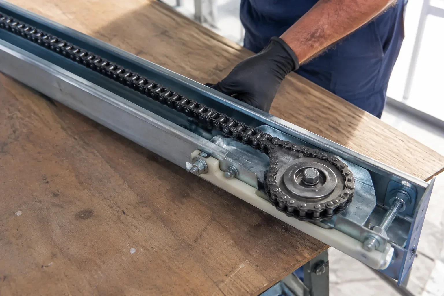 Gloved hand resting on a steel opener rail laid on a wooden bench, showing a drive chain looped around a sprocket wheel. — Chain-Drive Opener Repair, Toronto